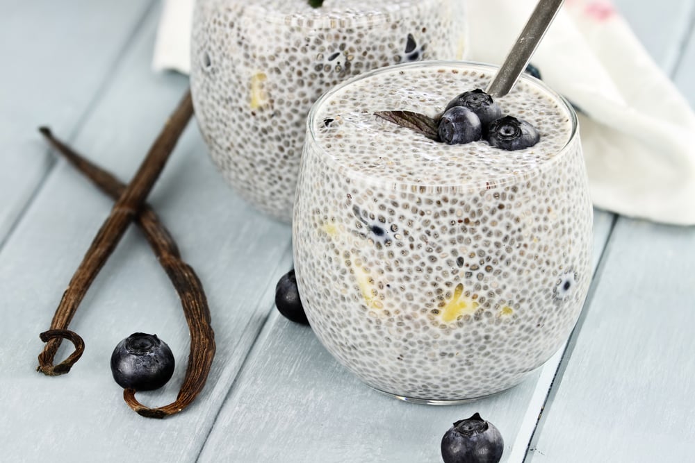 Chia pudding in a round glass with blueberries and bananas on a light blue picnic table by vanilla beans and blueberries