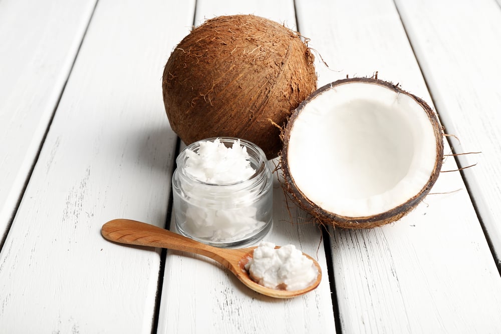 a halved coconut on a white picnic style table in front of a jar of coconut oil and a wooden spoon